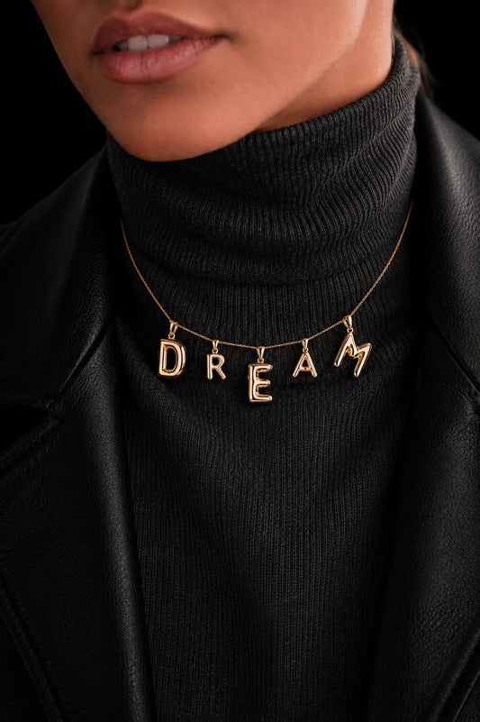 Gold necklace with 'DREAM' pendant on a black turtleneck sweater.