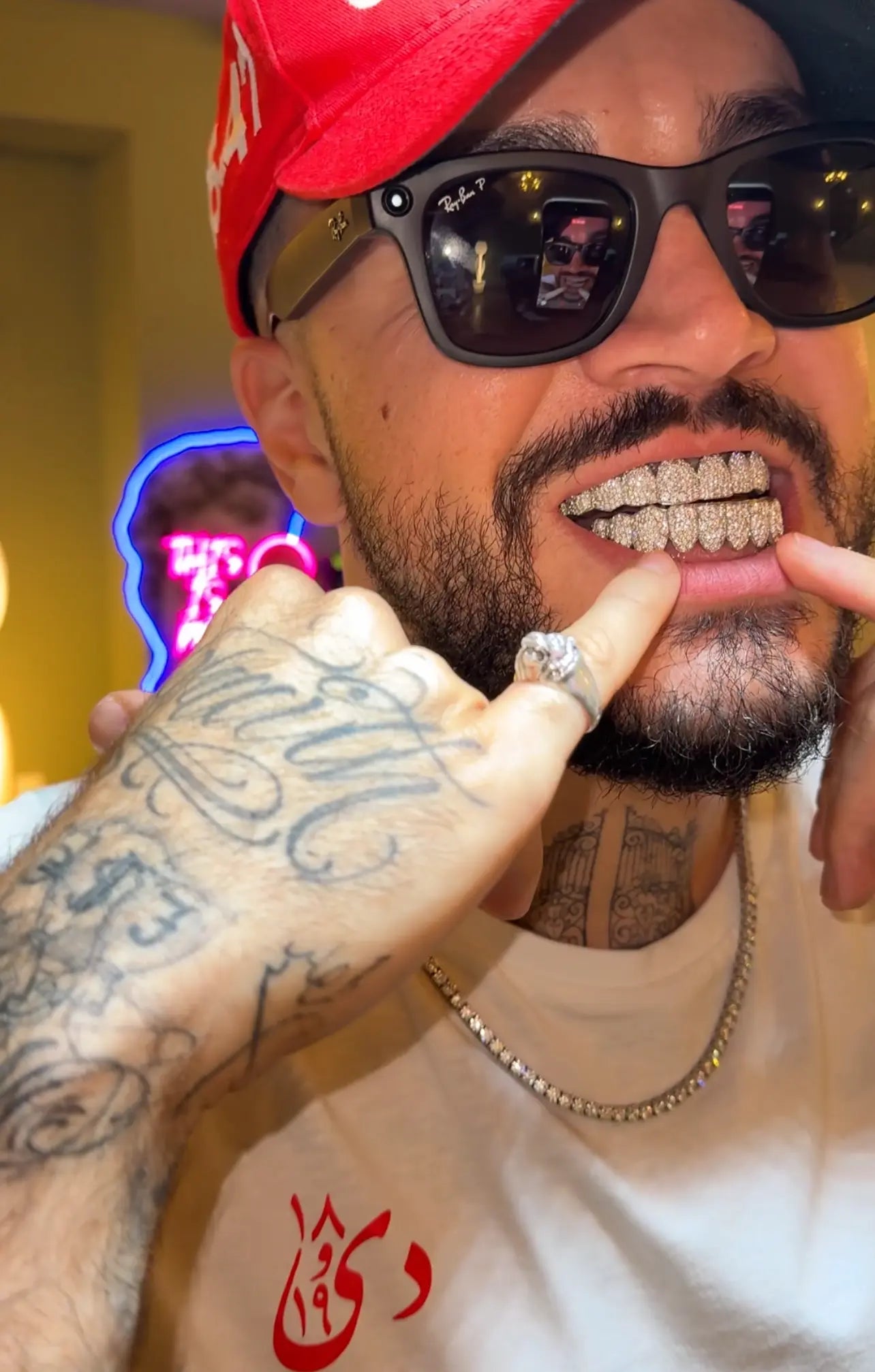 A close-up selfie of a man wearing a red baseball cap, black sunglasses, a white t-shirt, and a gold chain necklace, pointing to his teeth which are covered in sparkling diamond grillz. He has a tattooed hand and a ring on his finger.