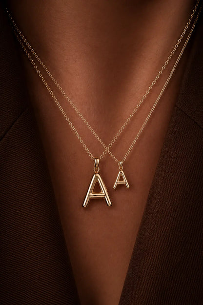 Two gold necklaces with 'A' pendants on a person's neck against a brown background.