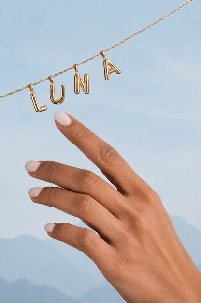 Gold necklace with 'LUNA' pendant held by a hand against a blue sky background