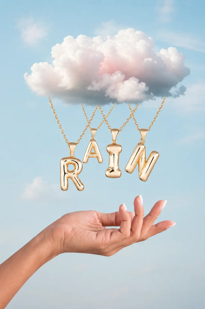 Gold necklace with 'RAIN' pendant held by a hand against a blue sky background