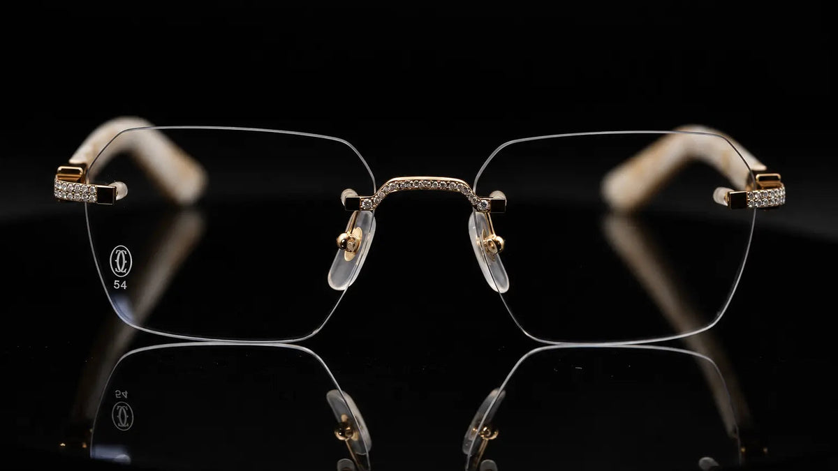 Rimless square eyeglasses with gold metal frames, crystal rhinestone accents on the temples, and clear nose pads, displayed on a reflective black surface against a black background.