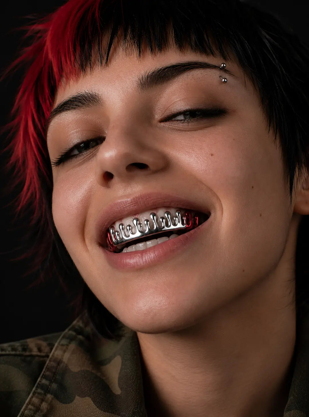 A close-up of a person with short black and bright red hair wearing silver metal grillz on their teeth, with a small stud piercing above the left eyebrow, set against a dark background.