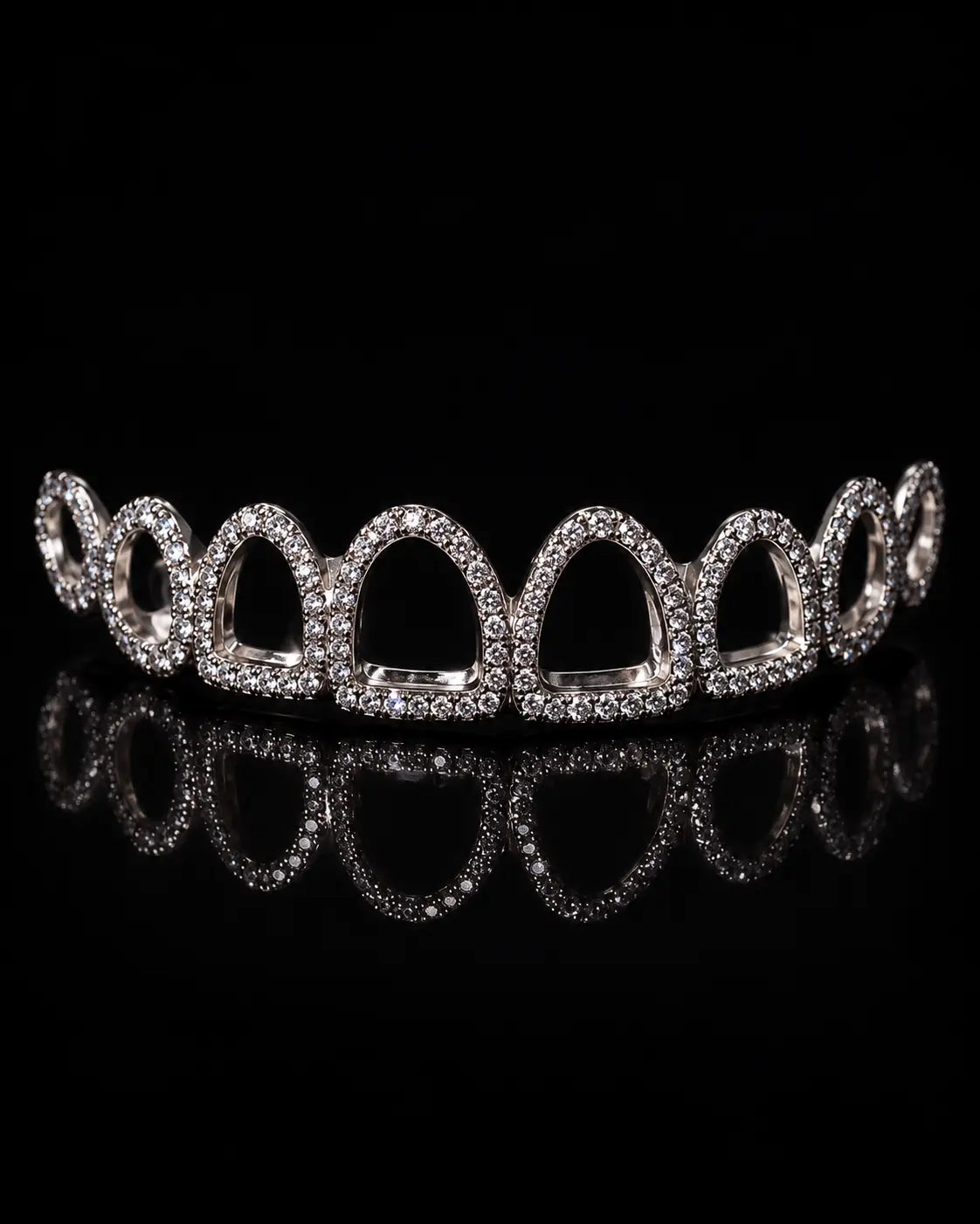A silver-toned arched crown or tiara embellished with clear pavé stones, displayed on a reflective black surface against a black background.