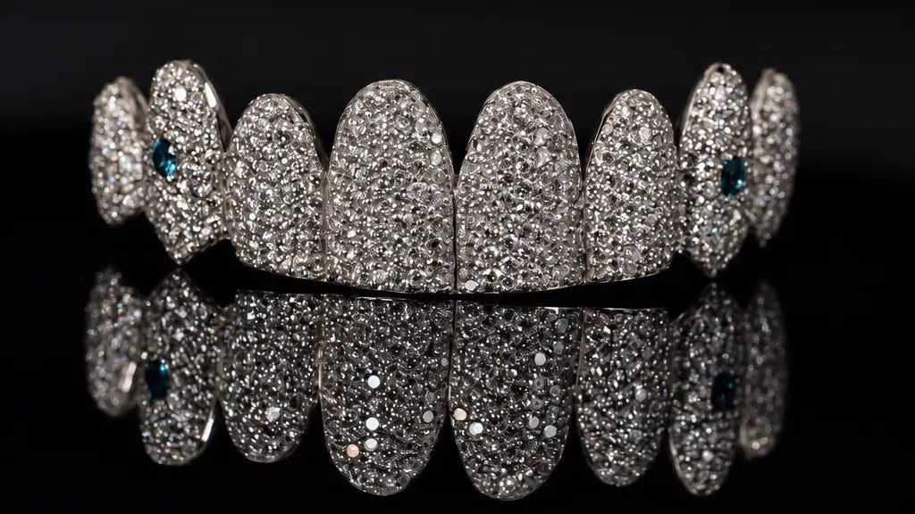 A row of custom teeth-shaped diamond grillz or mouthguards in silver-toned metal, heavily paved with clear diamonds, with tiny dark blue gemstone accents on the outer edges, displayed on a reflective black surface against a black background.