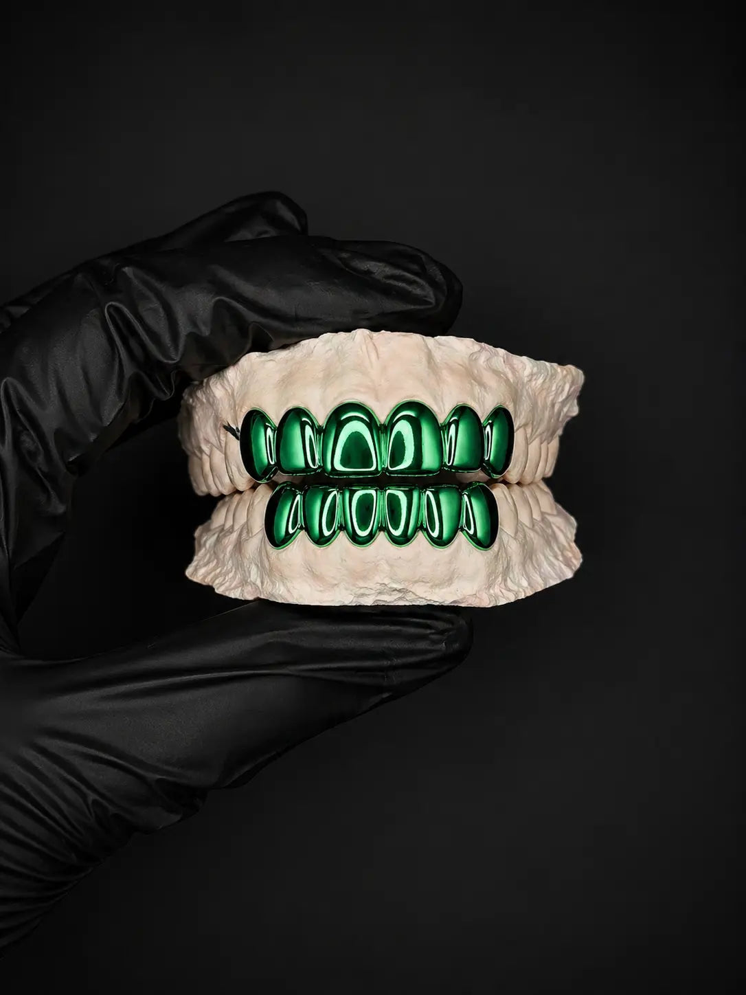 Grillz with green accents held by a gloved hand against a black background