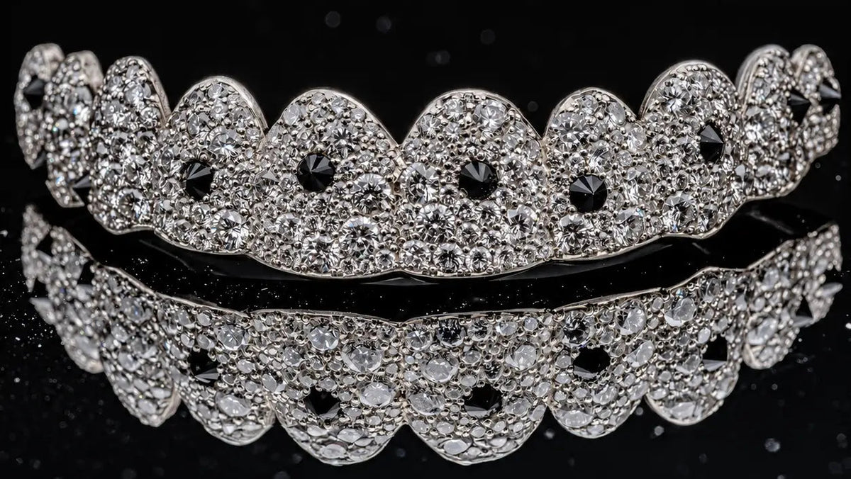 A close-up of a silver-toned dental grill or mouthguard with a scalloped edge, heavily embellished with clear pavé stones and accented with small black stones, set against a reflective black background.