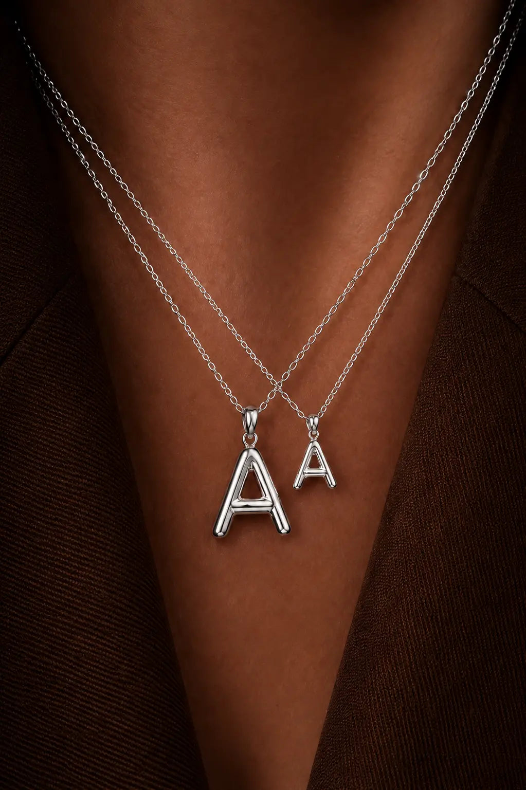 Two silver necklaces with 'A' pendants on a person's neck against a brown background.