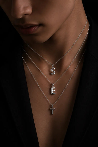 Close-up of a person wearing silver necklaces with 'S' and 'E' pendants on a dark background