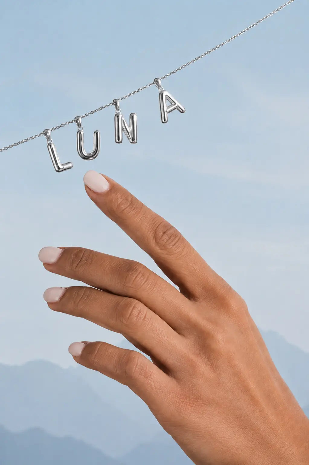Silver necklace with 'LUNA' pendant held by a hand against a blue sky.