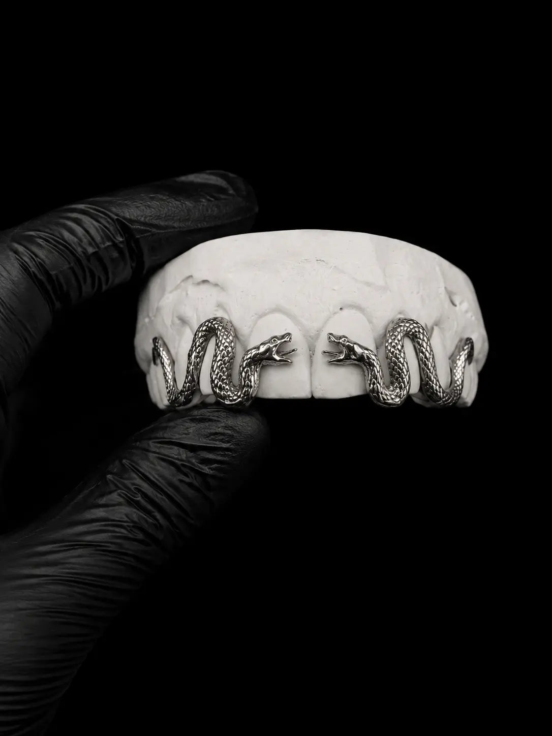 Gold snake grillz held by a gloved hand against a black background